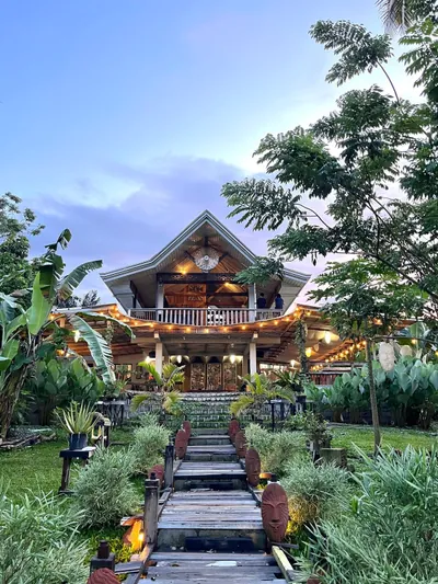 Alitaptap Art Cafe at dusk — a two-storey timber pavilion with a wide curved roof and warm string lights, at the end of a boardwalk lined with carved terracotta faces, banana plants and palms under a violet sky