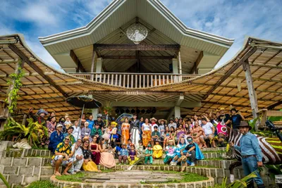 Dozens of people of all ages in costume, gathered for a group portrait on the steps of the same curved-roof pavilion under a bright blue sky